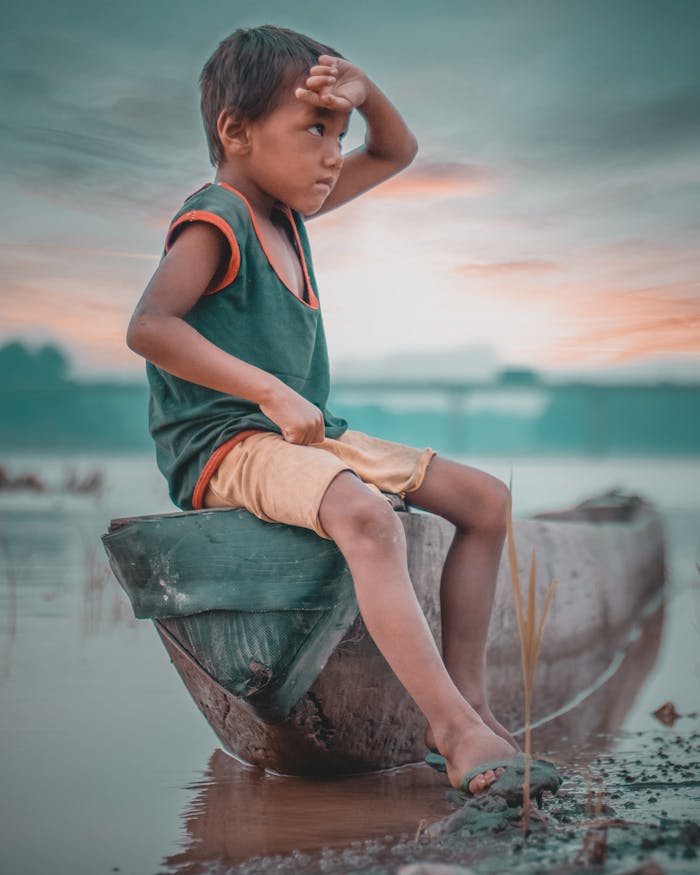 services-04 A young boy sits on a wooden boat by a misty riverside in Nepal, gazing into the distance.